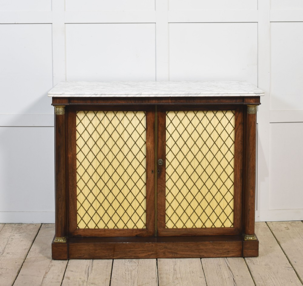 A nineteenth-century dark rosewood two-door cabinet featuring a white marble top, brass grille doors with pleated fabric lining, and columnar side supports.