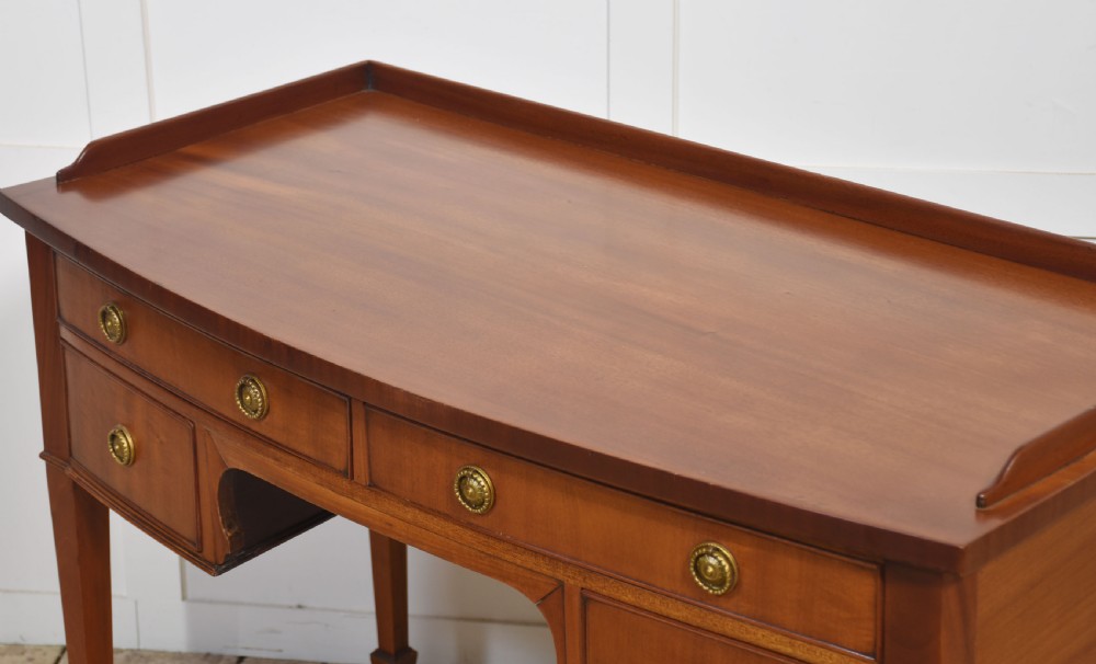 An antique warm brown mahogany writing table featuring a low gallery top, four drawers with circular brass pulls, tapered square legs, and brass castors. View V