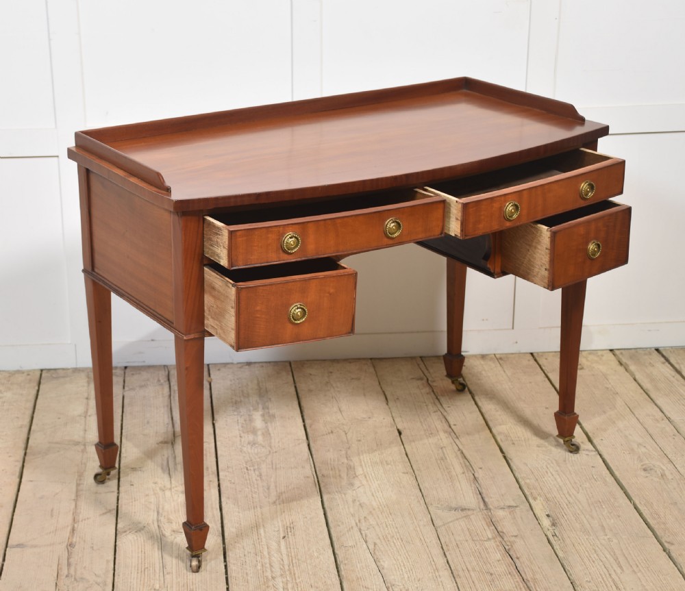 An antique warm brown mahogany writing table featuring a low gallery top, four drawers with circular brass pulls, tapered square legs, and brass castors. View XI