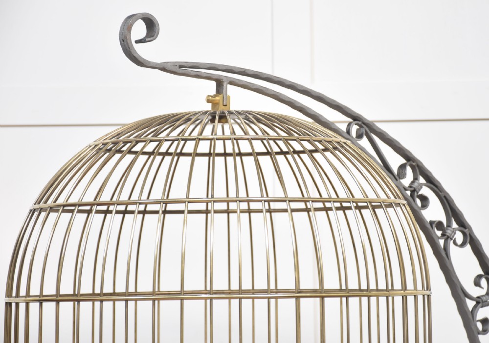 A domed brass bird cage with a copper base tray, suspended from a grey crescent-shaped wrought iron stand. View III