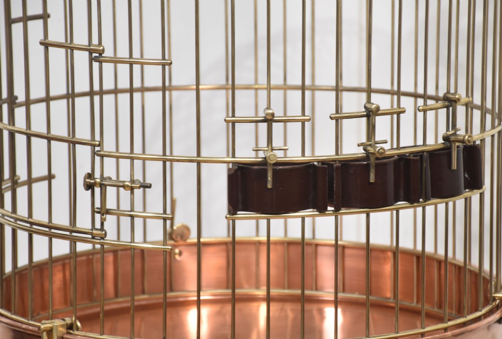 A domed brass bird cage with a copper base tray, suspended from a grey crescent-shaped wrought iron stand. View VII