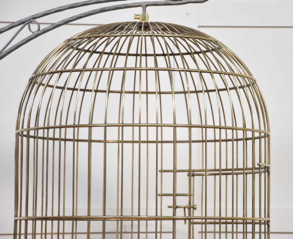 A domed brass bird cage with a copper base tray, suspended from a grey crescent-shaped wrought iron stand. View X