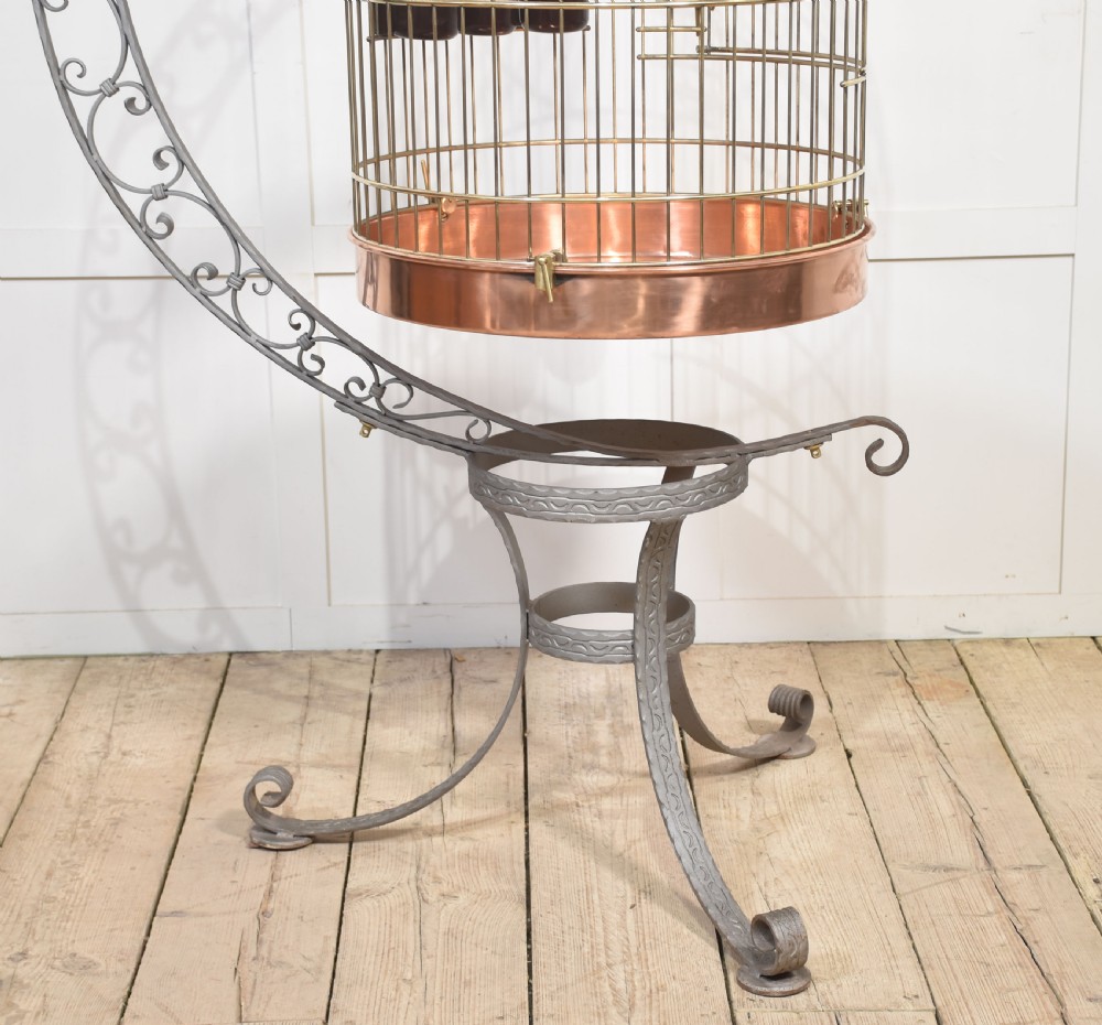 A domed brass bird cage with a copper base tray, suspended from a grey crescent-shaped wrought iron stand. View XII