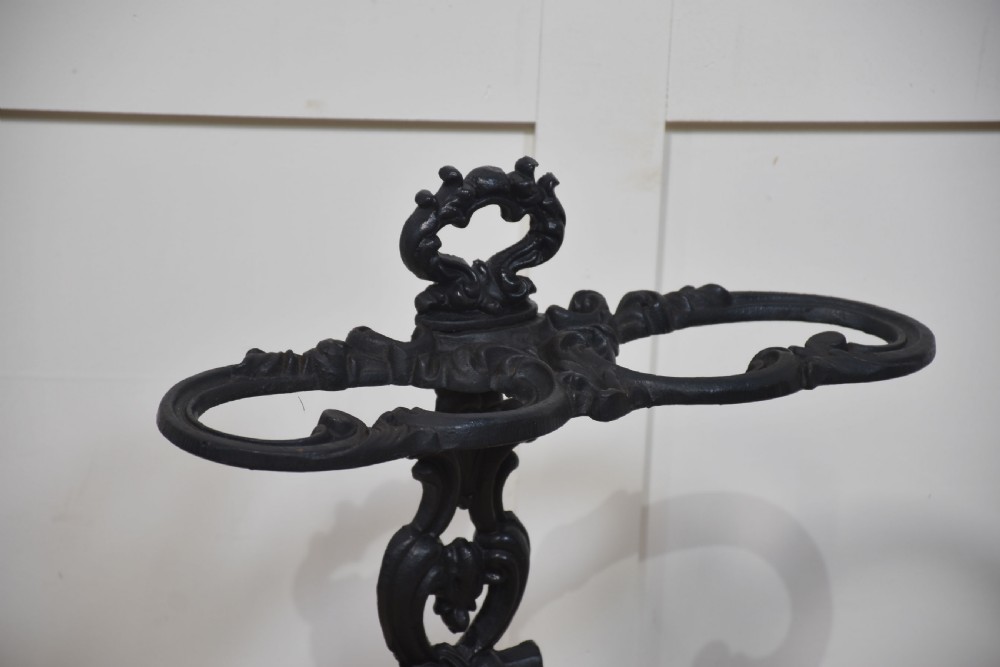 A black ornate cast iron umbrella stand featuring central scrollwork, an upper division ring, a top carrying loop, and a double-well drip tray base. View IV