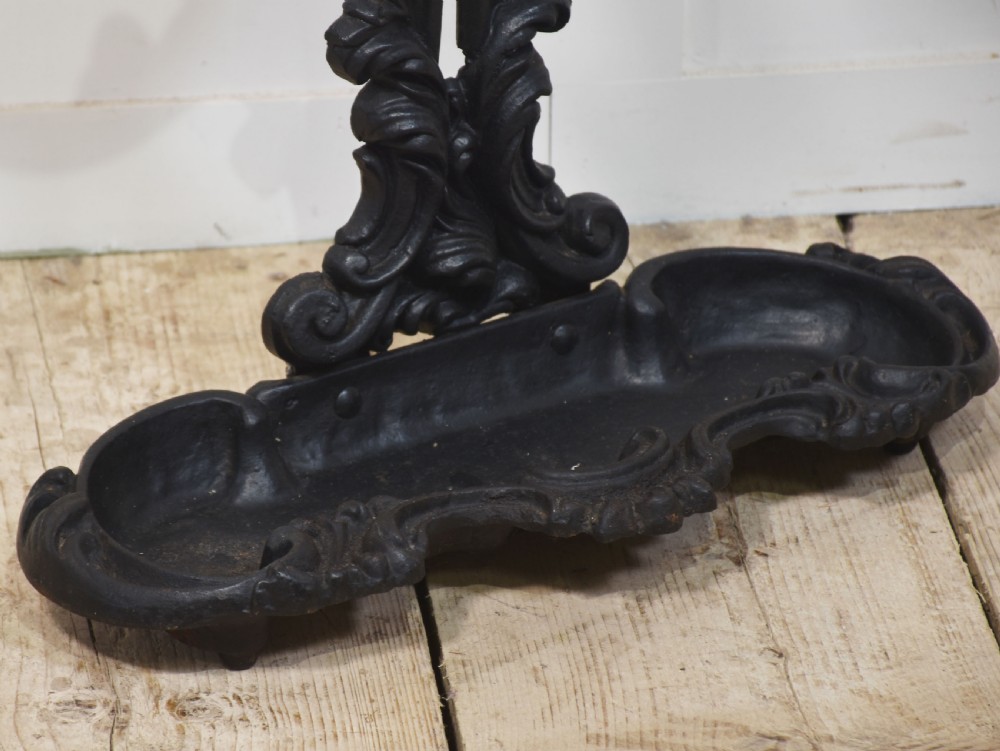A black ornate cast iron umbrella stand featuring central scrollwork, an upper division ring, a top carrying loop, and a double-well drip tray base. View VI