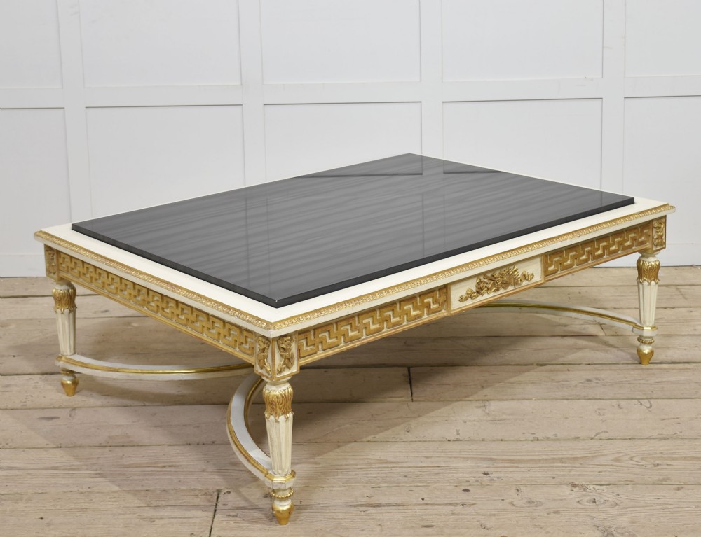 A low rectangular Venetian style coffee table featuring a black stone top, cream painted frieze with gold Greek key carving, and parcel gilt fluted legs.