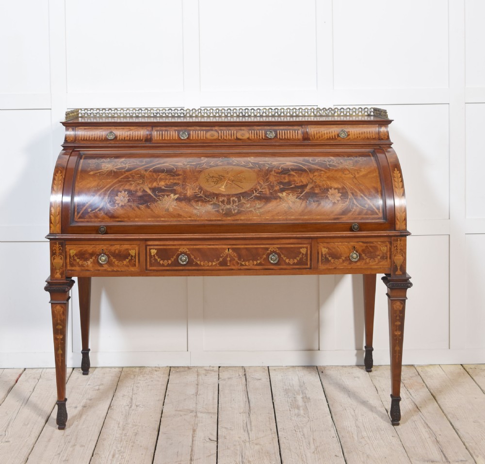 A Sheraton Revival mahogany cylinder bureau featuring intricate marquetry inlays on the curved roll-top lid, a brass gallery top, three drawers, and tapered legs.