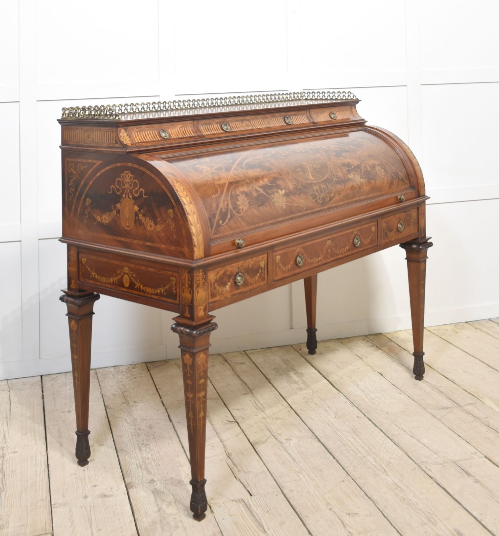 A Sheraton Revival mahogany cylinder bureau featuring intricate marquetry inlays on the curved roll-top lid, a brass gallery top, three drawers, and tapered legs. View VI