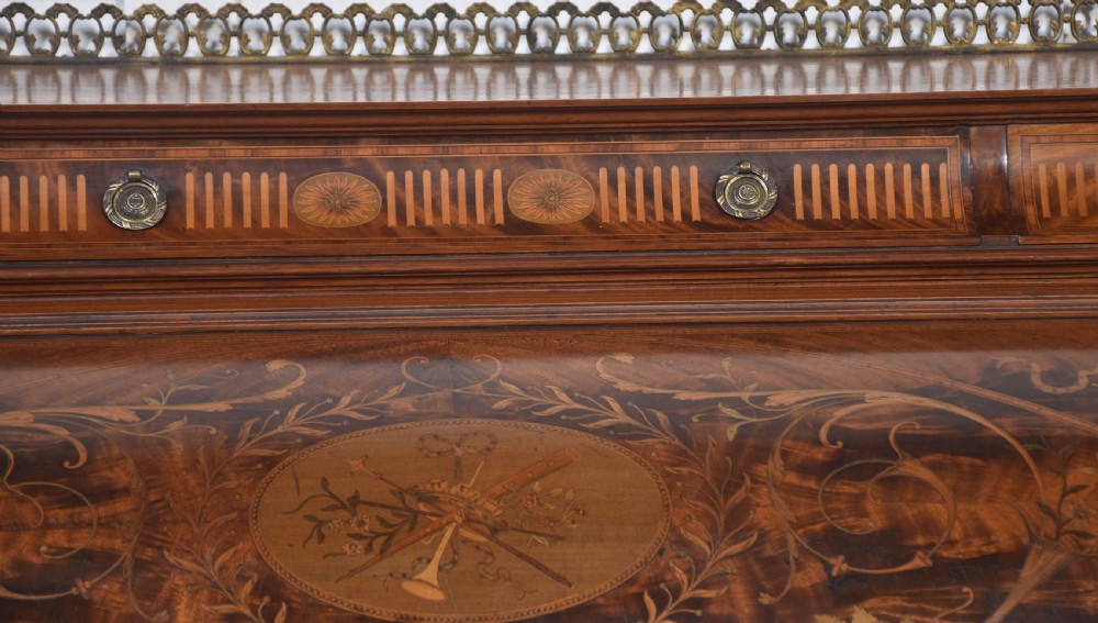 A Sheraton Revival mahogany cylinder bureau featuring intricate marquetry inlays on the curved roll-top lid, a brass gallery top, three drawers, and tapered legs. View VIII