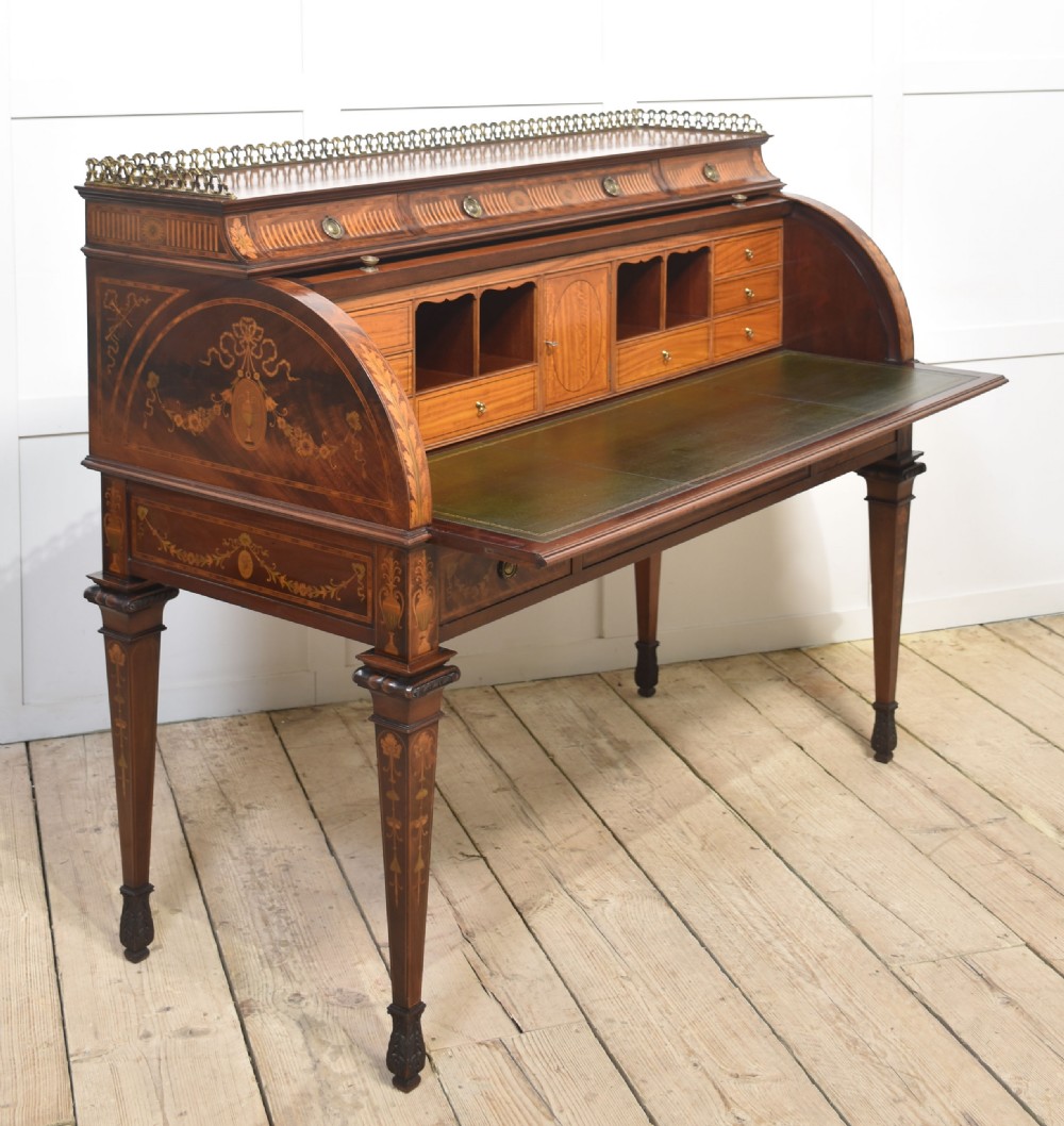 A Sheraton Revival mahogany cylinder bureau featuring intricate marquetry inlays on the curved roll-top lid, a brass gallery top, three drawers, and tapered legs. View XII