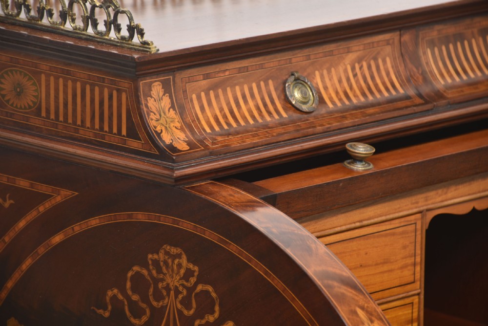 A Sheraton Revival mahogany cylinder bureau featuring intricate marquetry inlays on the curved roll-top lid, a brass gallery top, three drawers, and tapered legs. View XIV