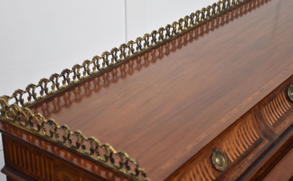 A Sheraton Revival mahogany cylinder bureau featuring intricate marquetry inlays on the curved roll-top lid, a brass gallery top, three drawers, and tapered legs. View XVI