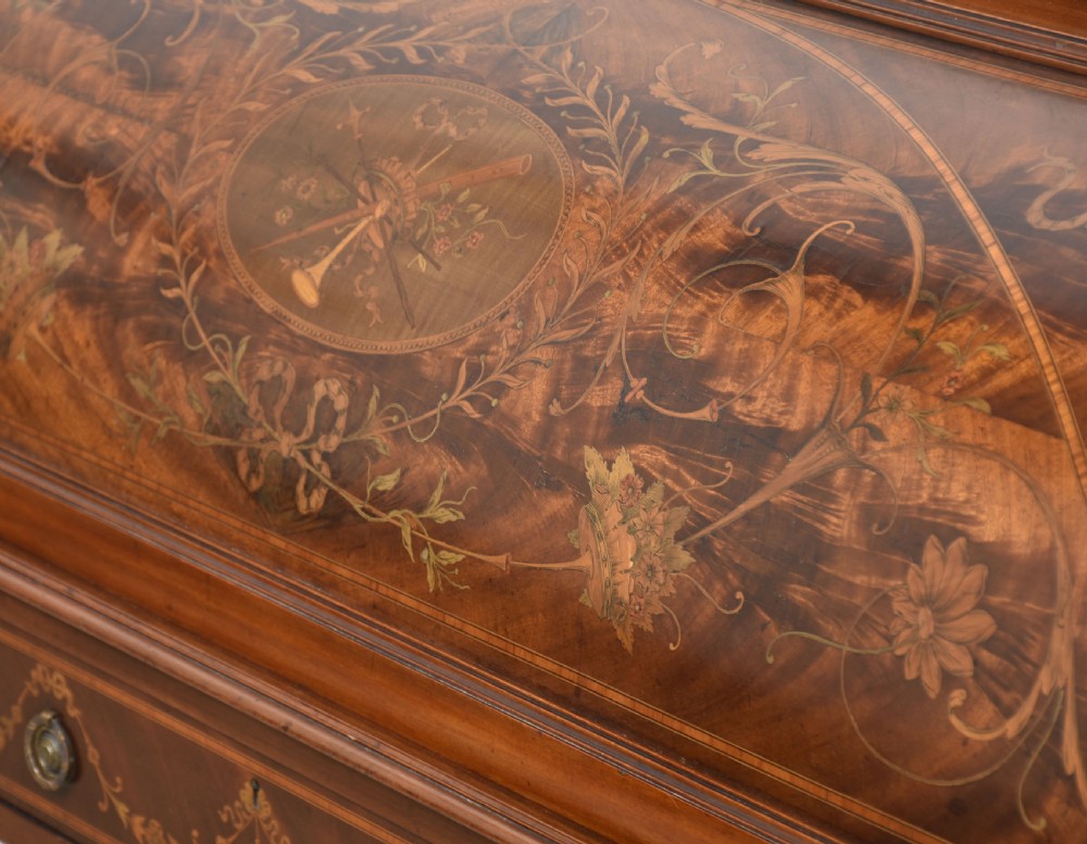 A Sheraton Revival mahogany cylinder bureau featuring intricate marquetry inlays on the curved roll-top lid, a brass gallery top, three drawers, and tapered legs. View XVII