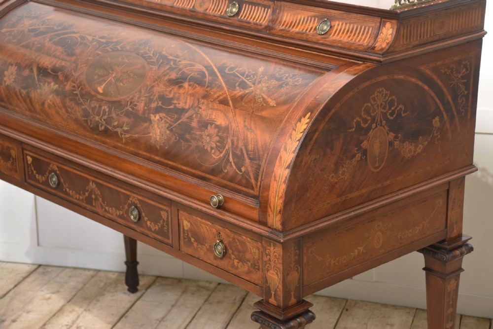 A Sheraton Revival mahogany cylinder bureau featuring intricate marquetry inlays on the curved roll-top lid, a brass gallery top, three drawers, and tapered legs. View XX