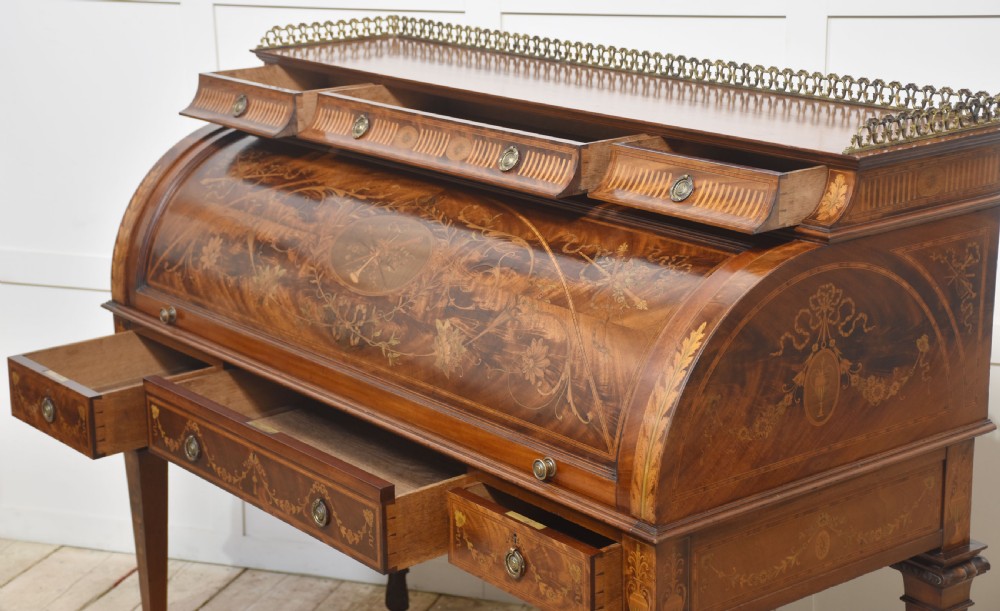 A Sheraton Revival mahogany cylinder bureau featuring intricate marquetry inlays on the curved roll-top lid, a brass gallery top, three drawers, and tapered legs. View XXI