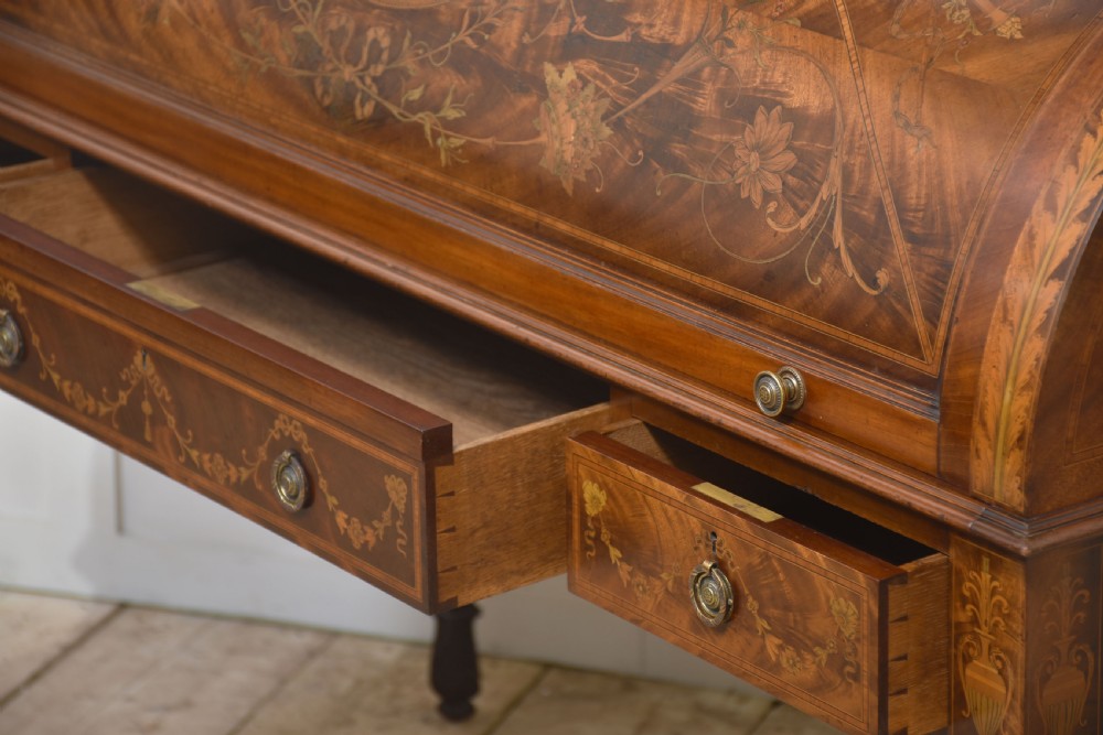 A Sheraton Revival mahogany cylinder bureau featuring intricate marquetry inlays on the curved roll-top lid, a brass gallery top, three drawers, and tapered legs. View XXII