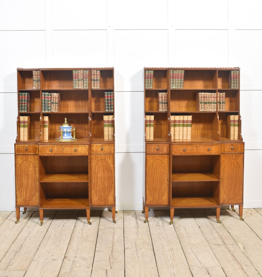 A matching pair of open satinwood breakfront bookcases featuring stepped waterfall shelving, inlaid drawers, and oval panel cupboard doors.