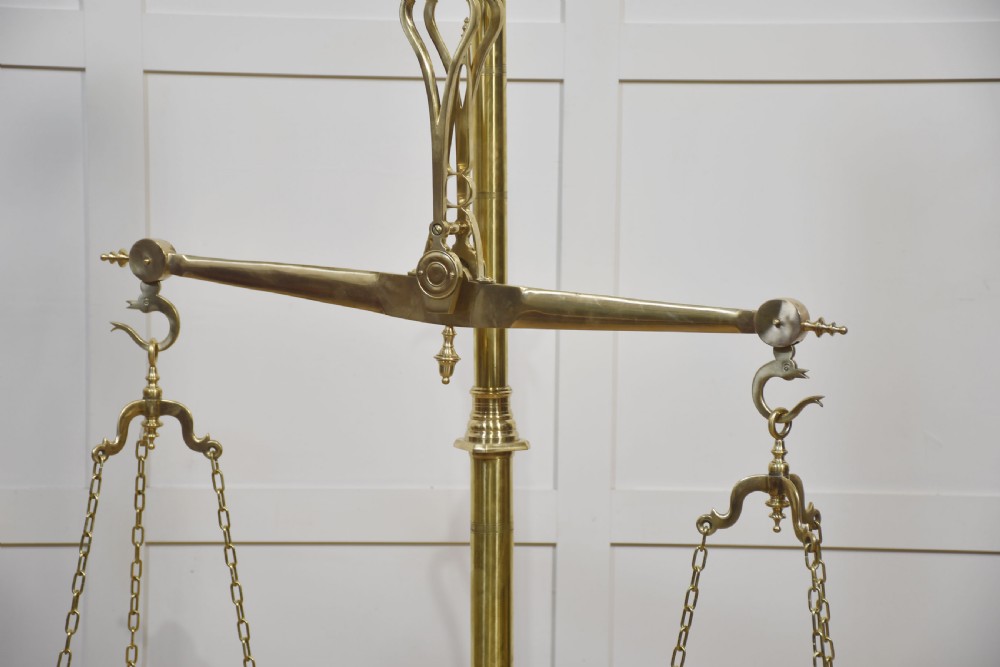 A floor-standing brass balance scale featuring a bull head finial, turned central column, horizontal beam, and two circular brass pans suspended by chains. View VI