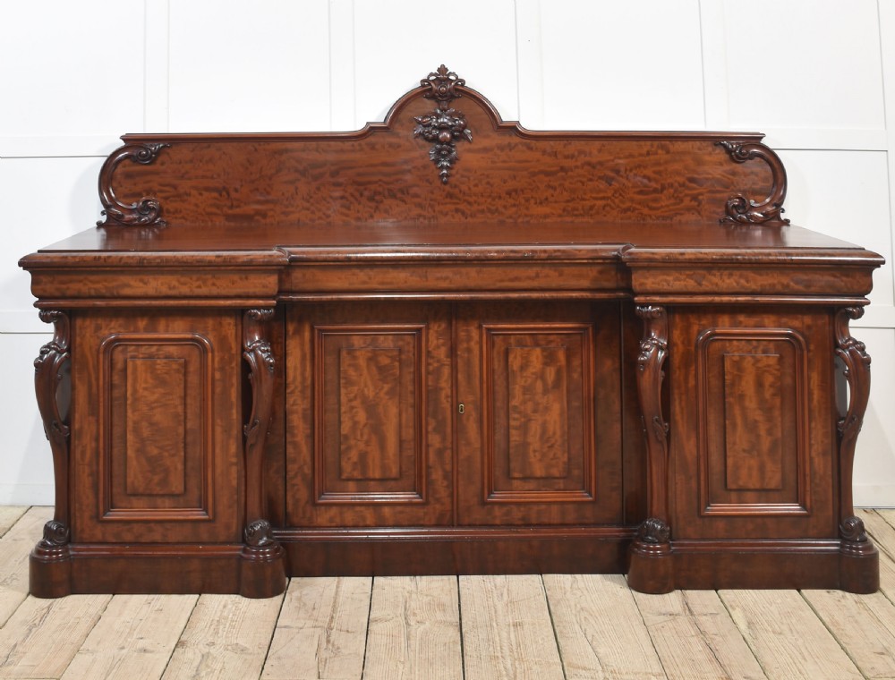 A large Victorian breakfront sideboard crafted from figured mahogany featuring four panelled doors, carved scroll brackets, and an arched cresting rail with carved floral motifs.