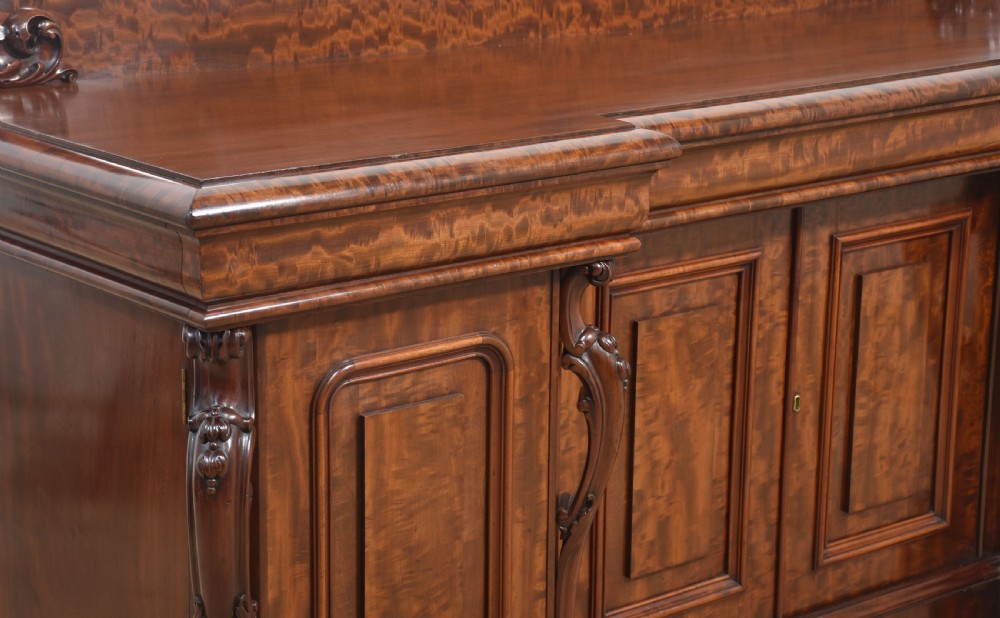 A large Victorian breakfront sideboard crafted from figured mahogany featuring four panelled doors, carved scroll brackets, and an arched cresting rail with carved floral motifs. View III