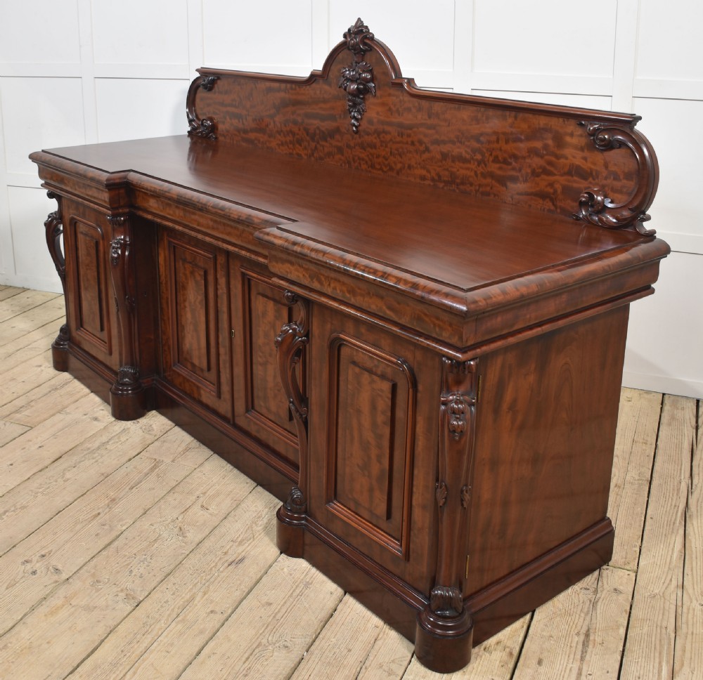 A large Victorian breakfront sideboard crafted from figured mahogany featuring four panelled doors, carved scroll brackets, and an arched cresting rail with carved floral motifs. View VI