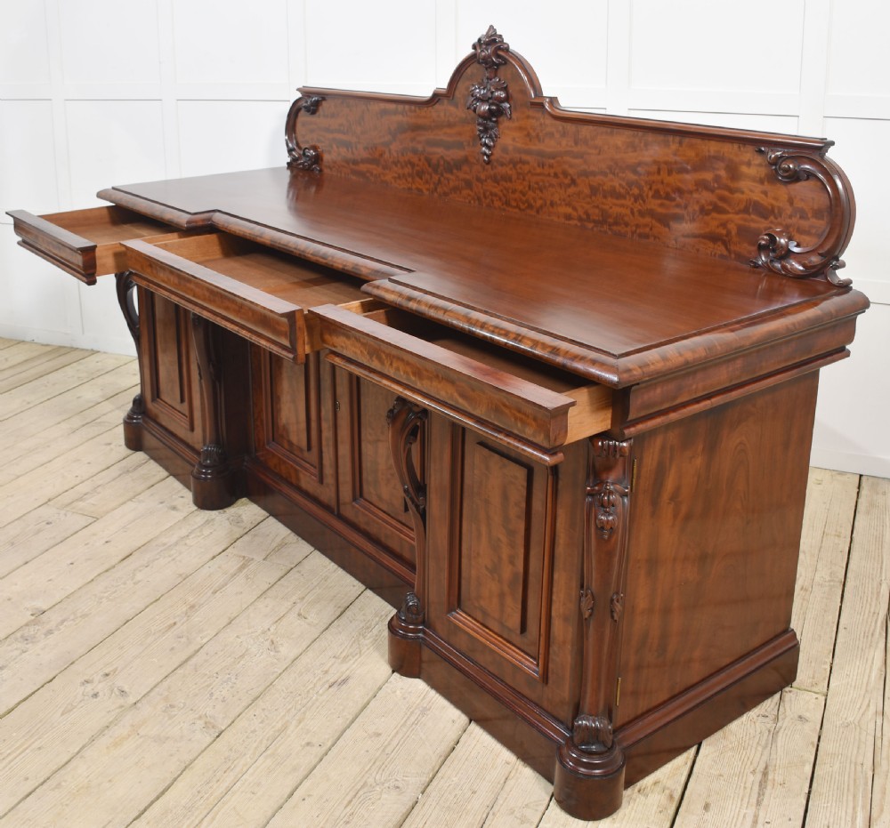 A large Victorian breakfront sideboard crafted from figured mahogany featuring four panelled doors, carved scroll brackets, and an arched cresting rail with carved floral motifs. View XI