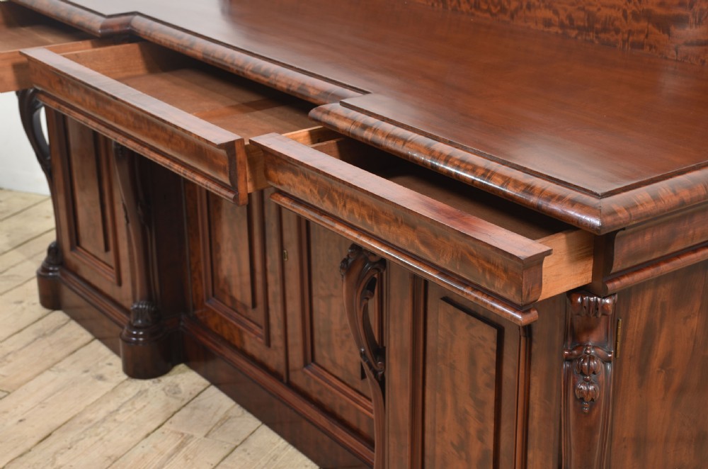 A large Victorian breakfront sideboard crafted from figured mahogany featuring four panelled doors, carved scroll brackets, and an arched cresting rail with carved floral motifs. View XII