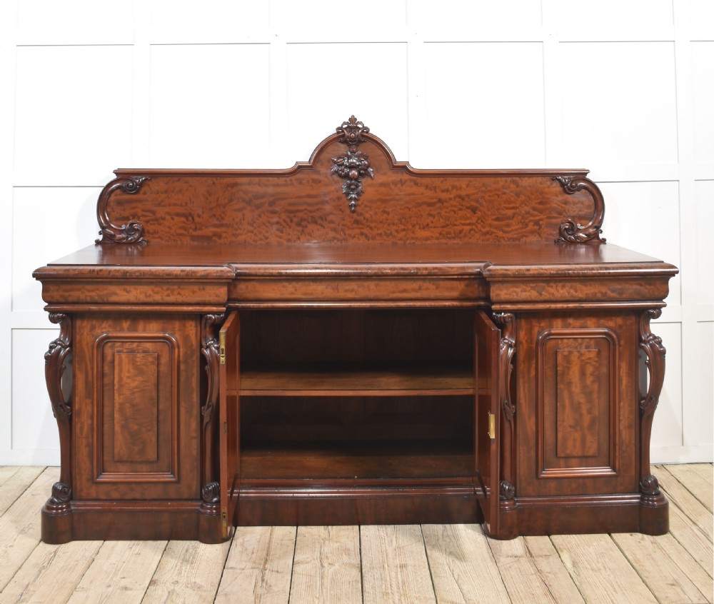 A large Victorian breakfront sideboard crafted from figured mahogany featuring four panelled doors, carved scroll brackets, and an arched cresting rail with carved floral motifs. View XVI