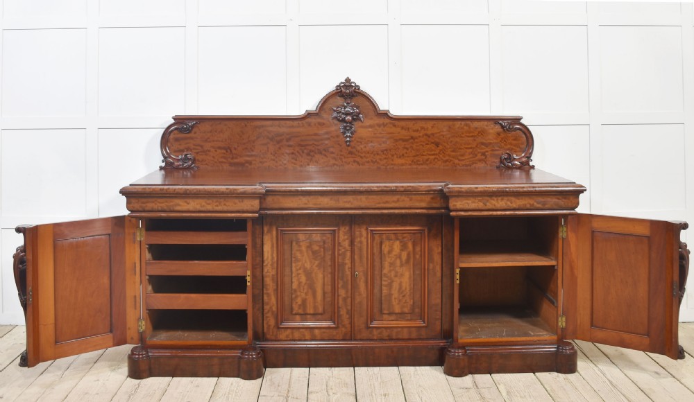 A large Victorian breakfront sideboard crafted from figured mahogany featuring four panelled doors, carved scroll brackets, and an arched cresting rail with carved floral motifs. View XVII