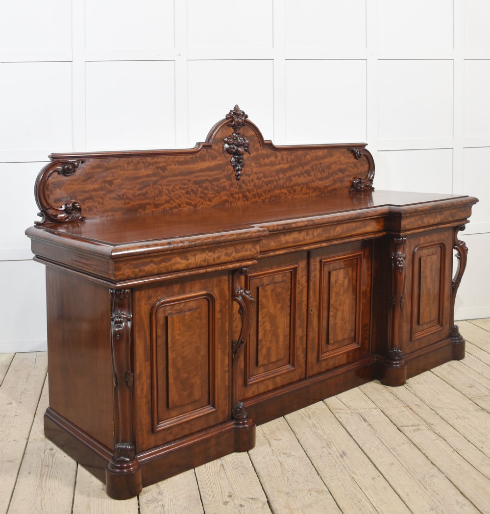 A large Victorian breakfront sideboard crafted from figured mahogany featuring four panelled doors, carved scroll brackets, and an arched cresting rail with carved floral motifs. View XVIII