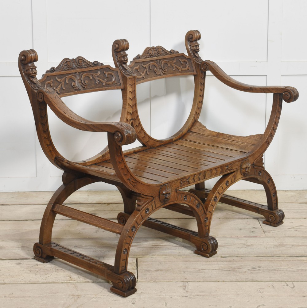 A carved oak double curule bench featuring scrolled armrests, carved mask details, and a slatted seat.