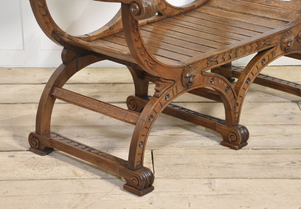 A carved oak double curule bench featuring scrolled armrests, carved mask details, and a slatted seat. View VI