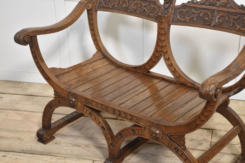 A carved oak double curule bench featuring scrolled armrests, carved mask details, and a slatted seat. View IX