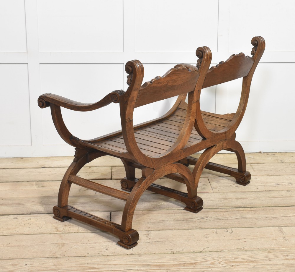 A carved oak double curule bench featuring scrolled armrests, carved mask details, and a slatted seat. View X