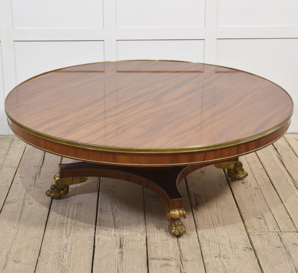 A circular mahogany coffee table featuring a polished flame mahogany veneer top, a thin brass gallery rim, and four curved legs ending in brass lion paw feet.