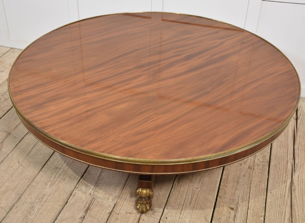 A circular mahogany coffee table featuring a polished flame mahogany veneer top, a thin brass gallery rim, and four curved legs ending in brass lion paw feet. View IV
