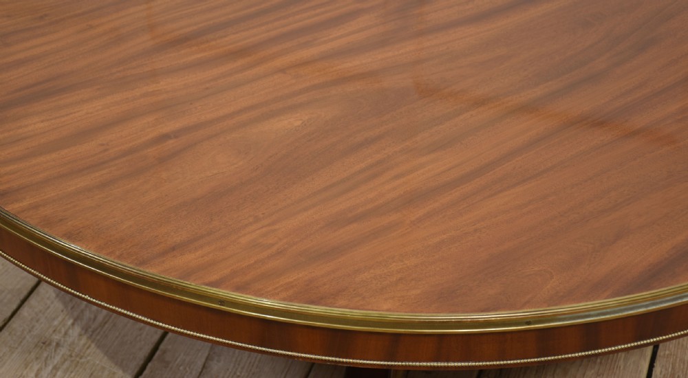 A circular mahogany coffee table featuring a polished flame mahogany veneer top, a thin brass gallery rim, and four curved legs ending in brass lion paw feet. View VII