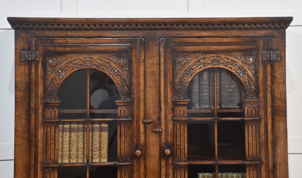 A tall dark oak two-door glazed bookcase featuring carved arched upper sections and panelled cupboard doors below. View II
