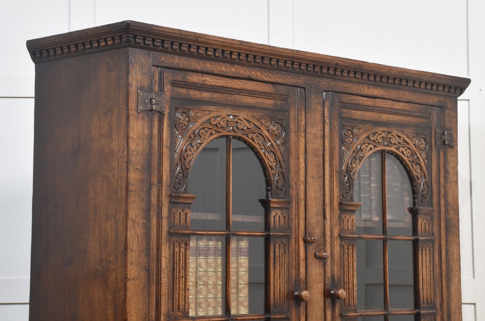 A tall dark oak two-door glazed bookcase featuring carved arched upper sections and panelled cupboard doors below. View X