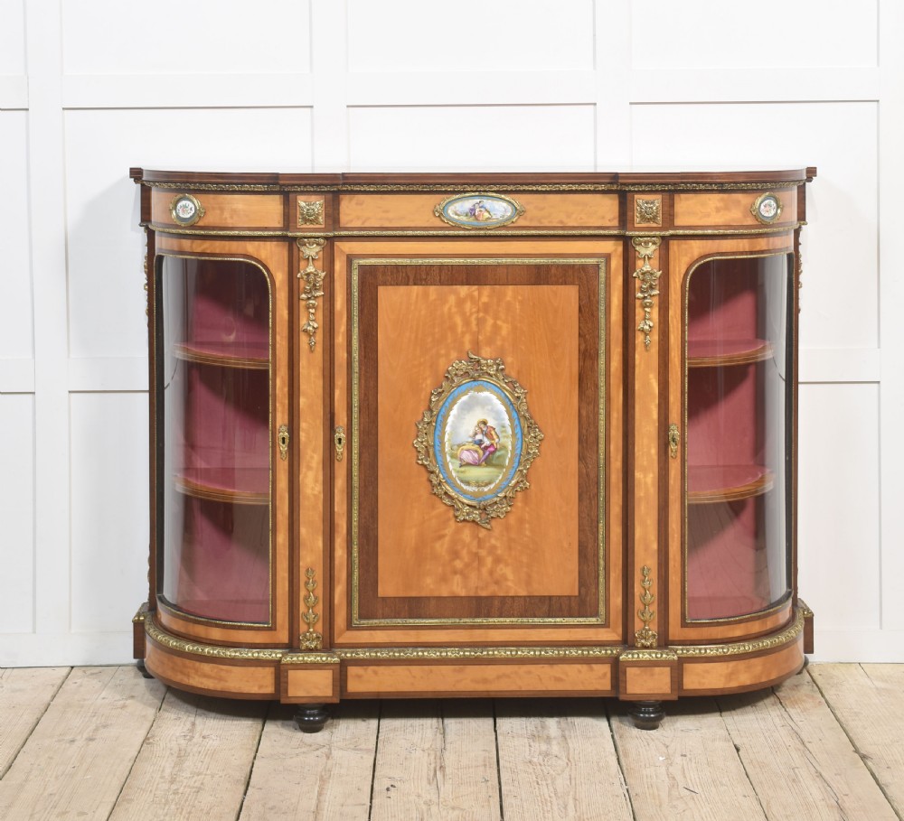 A 19th-century satinwood credenza featuring curved glass side display doors, a central solid panel door set with a porcelain plaque, and gilded ormolu mounts throughout.