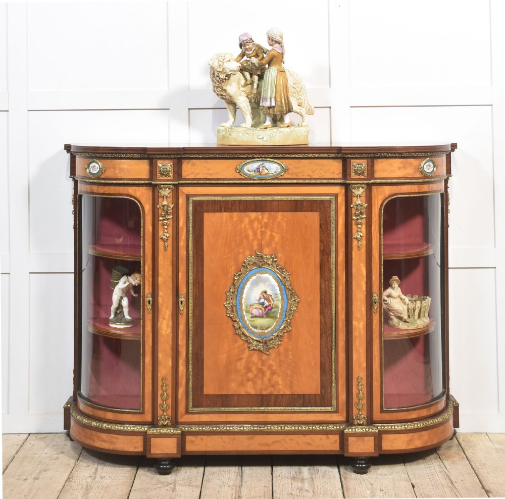 A 19th-century satinwood credenza featuring curved glass side display doors, a central solid panel door set with a porcelain plaque, and gilded ormolu mounts throughout. View II