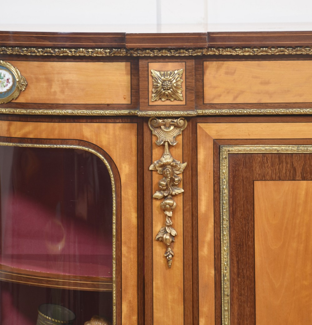 A 19th-century satinwood credenza featuring curved glass side display doors, a central solid panel door set with a porcelain plaque, and gilded ormolu mounts throughout. View V