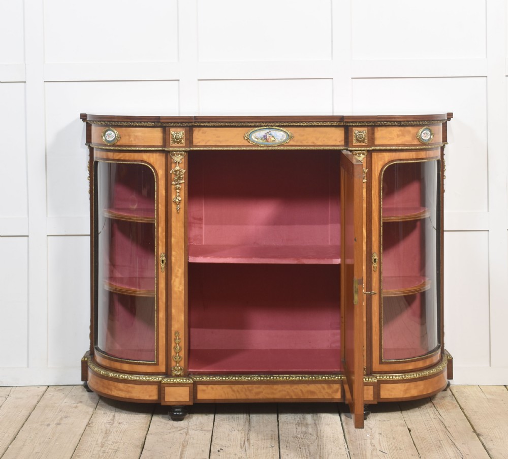 A 19th-century satinwood credenza featuring curved glass side display doors, a central solid panel door set with a porcelain plaque, and gilded ormolu mounts throughout. View VII