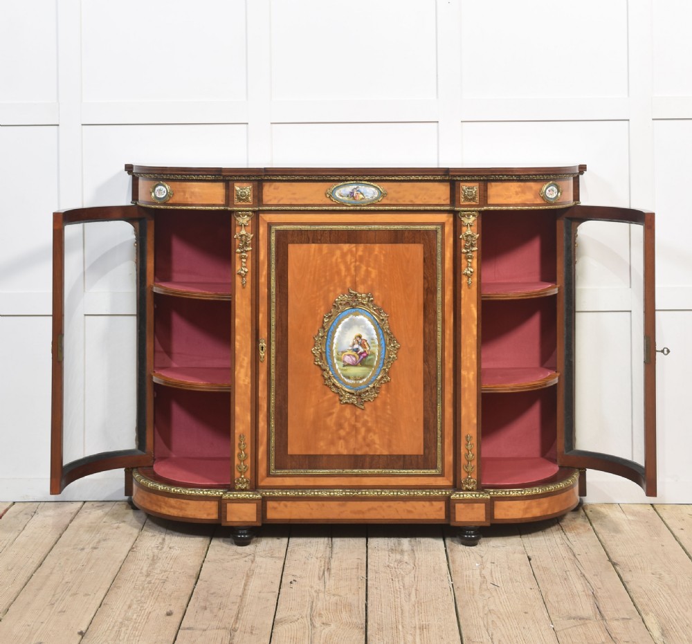 A 19th-century satinwood credenza featuring curved glass side display doors, a central solid panel door set with a porcelain plaque, and gilded ormolu mounts throughout. View VIII