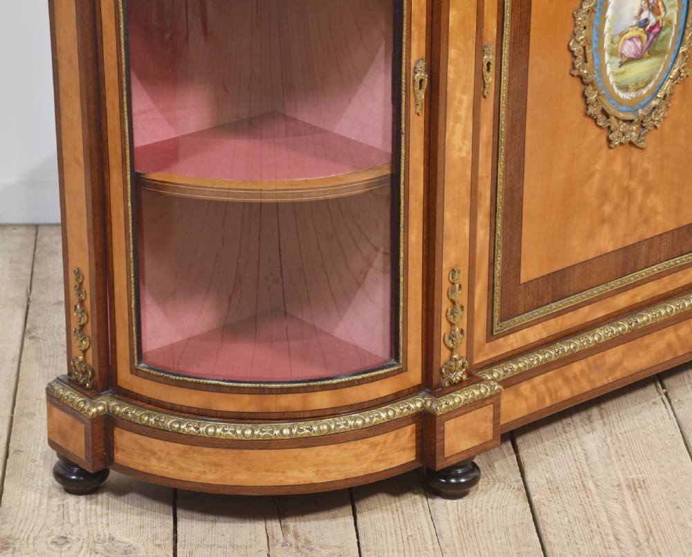 A 19th-century satinwood credenza featuring curved glass side display doors, a central solid panel door set with a porcelain plaque, and gilded ormolu mounts throughout. View X
