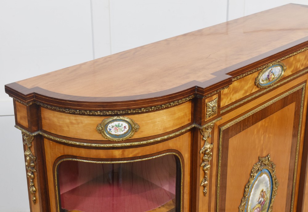 A 19th-century satinwood credenza featuring curved glass side display doors, a central solid panel door set with a porcelain plaque, and gilded ormolu mounts throughout. View XI