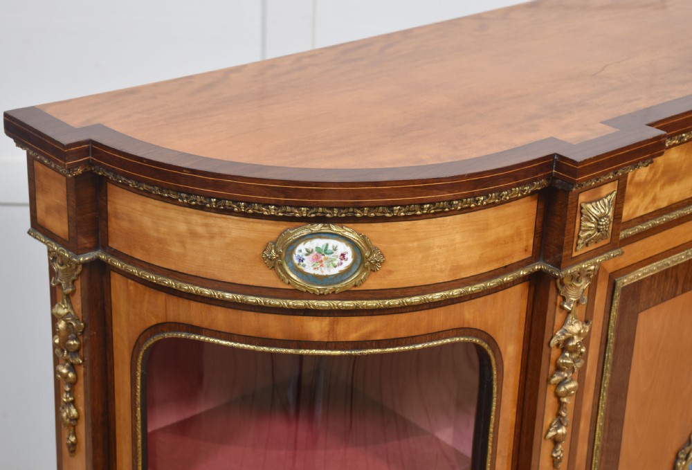 A 19th-century satinwood credenza featuring curved glass side display doors, a central solid panel door set with a porcelain plaque, and gilded ormolu mounts throughout. View XII