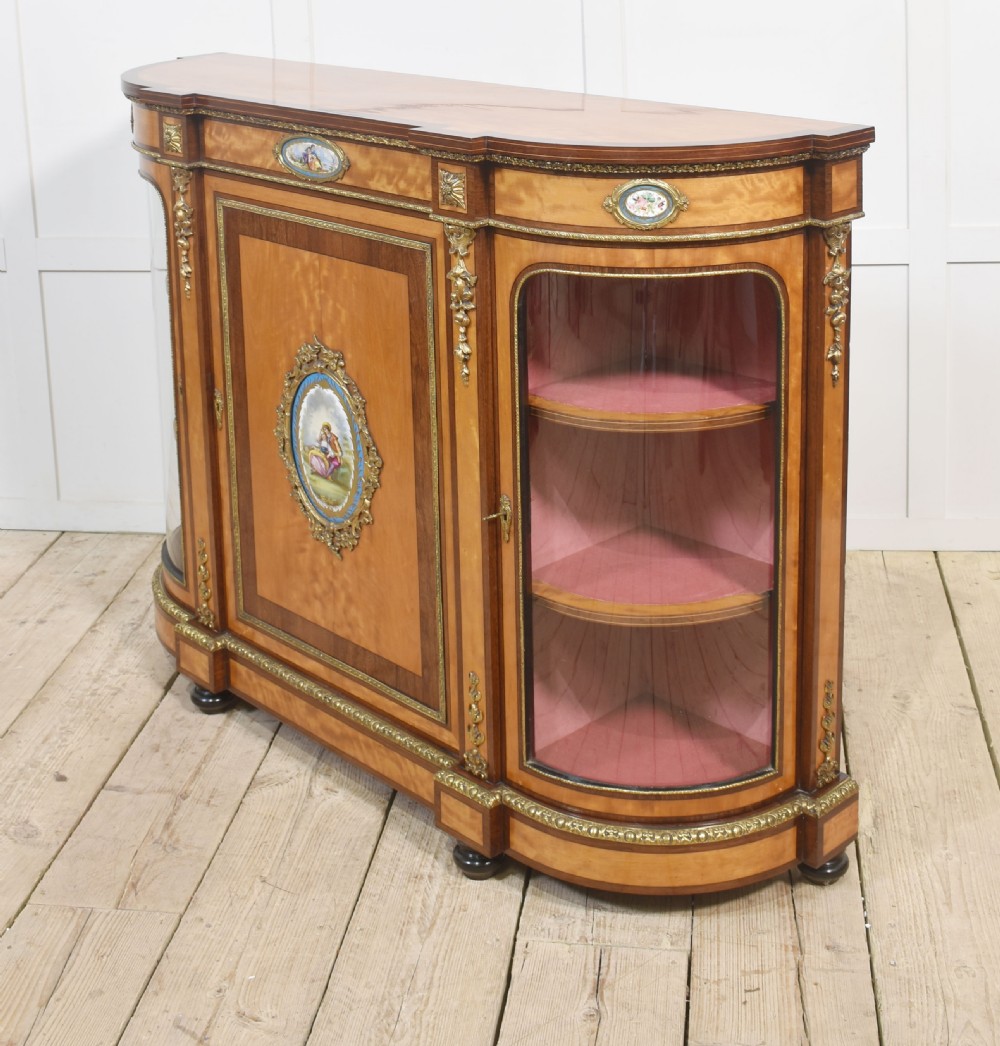 A 19th-century satinwood credenza featuring curved glass side display doors, a central solid panel door set with a porcelain plaque, and gilded ormolu mounts throughout. View XIII