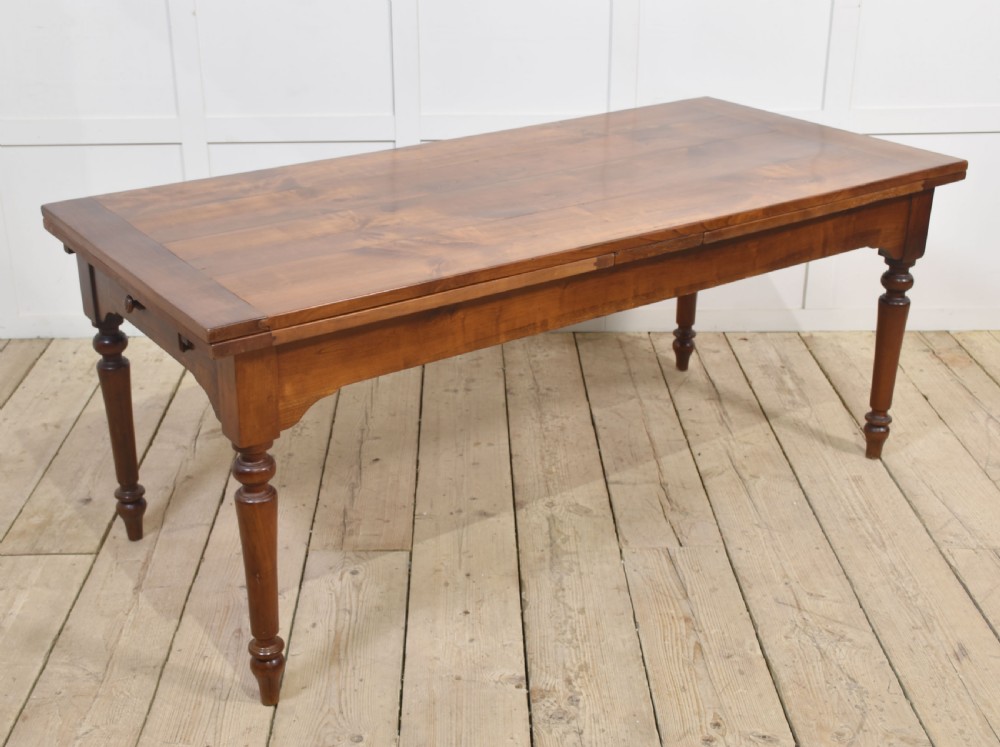 A large antique fruitwood farmhouse dining table featuring a smooth planked top, straight apron, and four turned legs resting on exposed wooden floorboards.