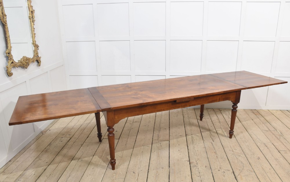 A large antique fruitwood farmhouse dining table featuring a smooth planked top, straight apron, and four turned legs resting on exposed wooden floorboards. View II